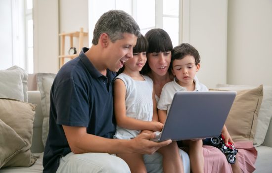 Excited cheerful parents couple holding kids on lap, sitting on couch all together, watching movie or video on laptop at home, staring at display. Medium shot. Communication or entertainment concept
