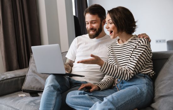 Happy young white couple looking at laptop computer sitting on a couch at home pointing finger
